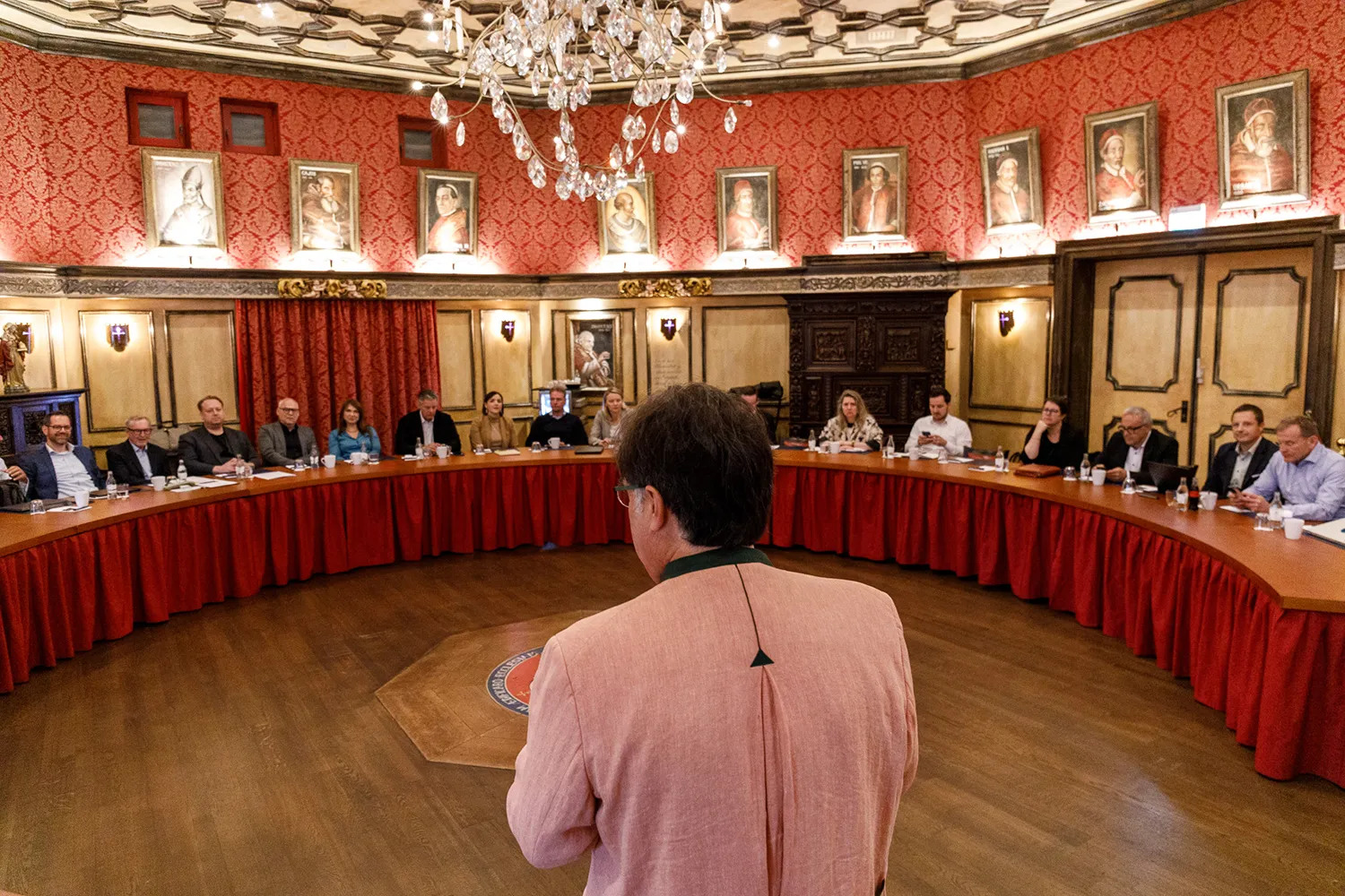 Runder Konferenzraum mit einer gebogenen Tischreihe an denen Personen sitzen und einer Person stehend in der Mitte